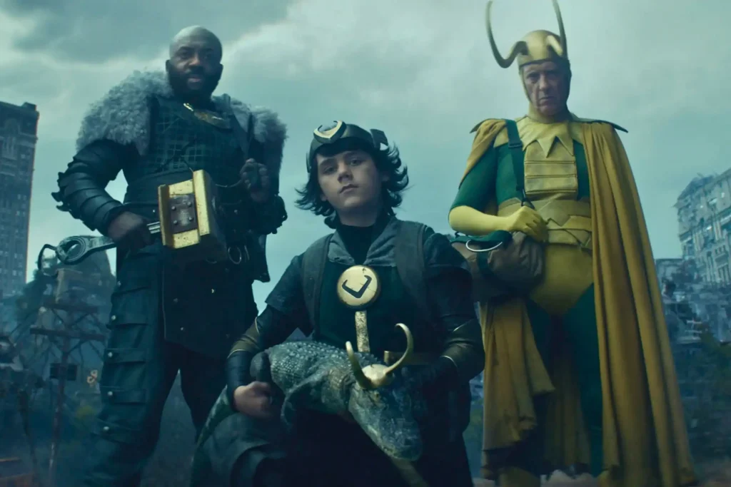 A full-body shot of three Loki variants and Alligator Loki standing in the desolate environment of The Void from the Marvel series Loki. From left to right: Boastful Loki (Deobia Oparei) in armor holding a small hammer, Kid Loki (Jack Veal) kneeling and holding Alligator Loki, and Classic Loki (Richard E. Grant) in his bright yellow and green comic-accurate costume. The background is a ruined city under a stormy sky.