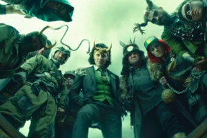A low-angle shot of several Loki variants from the MCU series Loki looking down, including the main Loki (Tom Hiddleston) in a green suit, Classic Loki, Kid Loki, Boastful Loki, and President Loki. They are standing in The Void, a desolate area at the end of time.