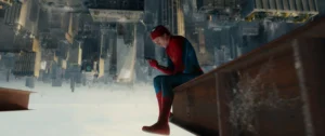 Spider-Man In Red And Blue Suit With Gold Spider Emblem Sitting On A Rooftop Ledge Looking At A Phone With New York Skyscrapers Behind Him